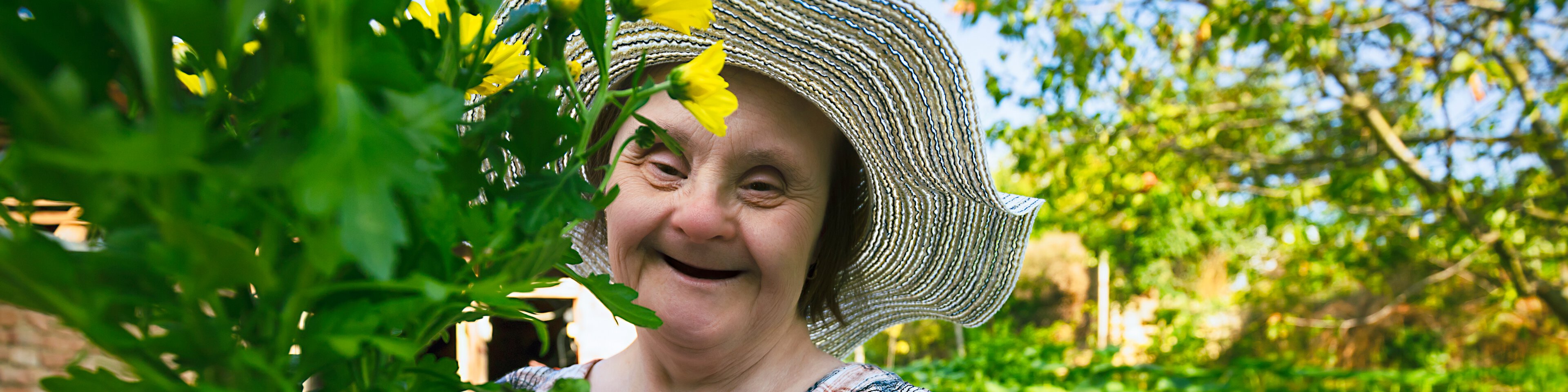 Eine ältere Frau mit Sonnenhut und Behinderung hält eine Pflanze in der Hand und lacht | © portishead1 - Getty Images